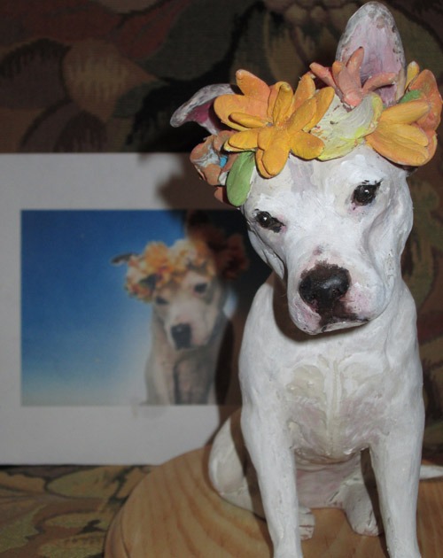 A handcrafted white dog figurine wears a vibrant flower crown, positioned in front of a framed photo of the same dog, highlighting a sentimental keepsake.